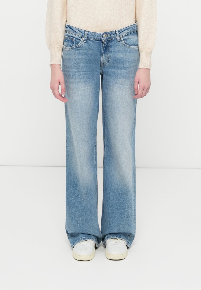 Svjetloplave široke traperice s klasičnim pet džepovima, s blagim izblijedjenjem na nogavicama. Uparene s bijelim tenisicama.