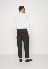 Schwarze maßgeschneiderte Hose mit lockerer Passform, elastischem Bund, zwei hinteren Taschen, kombiniert mit einem weißen Hemd und schwarzen Schuhen.