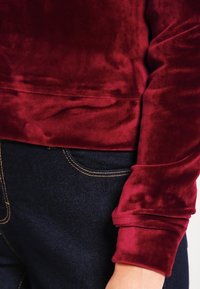 Top de terciopelo burdeos con una textura suave, que presenta un diseño ajustado y mangas con puños, combinado con jeans de mezclilla oscura.