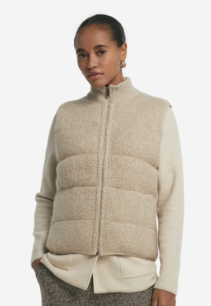 Beige vadderad väst med hög krage och full dragkedja, med en texturerad yttertyg och mjukt foder, lagrad över en lätt tröja.
