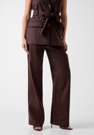 Mujer con pantalones anchos de color marrón oscuro y chaleco sin mangas con cinturón y bolsillos con solapa, combinado con sandalias de tacón abierto.
