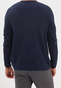 Muž ve námořnickém modrém pleteném svetr a šedých kostkovaných kalhotách, zobrazený zezadu na jednoduchém pozadí.