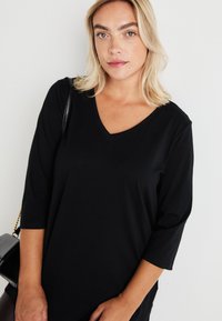 Črn top z V-izrezom in tromestnimi rokavi, narejen iz gladkega in raztegljivega materiala. Ima sproščen kroj in minimalističen dizajn.