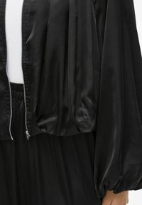 Černá bomber bunda z atlazového materiálu s předním zipem, nafouklými rukávy a volnějším střihem. Hladká textura s mírným leskem. Designové detaily na míru.