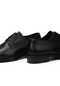 Zapatos de vestir de cuero negro con textura suave, punta redondeada y costuras sutiles. Presentan un diseño de cordones y una suela de goma de perfil bajo.