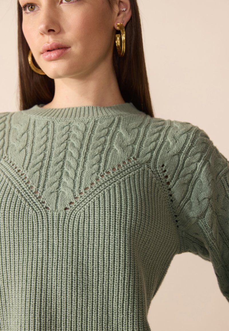 Maglione verde menta lavorato a maglia con un motivo a trecce, coste testurizzate e cuciture decorative lungo il colletto e le spalle.