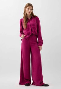 Camicia button-up in satin fucsia con due tasche, abbinata a pantaloni a gamba larga coordinati. Texture liscia e vestibilità ampia. Calzature nere.