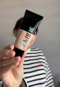 Fond de teint Maybelline Fit Me Matte et Poreless dans un tube de 30 ml, avec un capuchon noir et un corps beige. L'étiquette inclut des informations sur l'IP et la teinte.