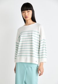 Pulover alb cu dungi orizontale albastre deschise, mâneci largi și tiv cu nervuri. Asortat cu pantaloni albastre deschise, pe un fundal simplu.