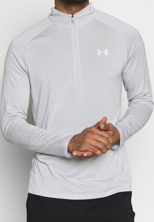 Mężczyzna ubrany w jasnoszarą sportową koszulkę z długim rękawem i zamkiem do ćwiartki, z logo Under Armour, z rękami złączonymi, na jednolitym tle.