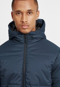 Jachetă puffer de culoare albastru marin cu fermoar în față, glugă și textură matlasată; prezintă un exterior neted și un design izolat pentru căldură.
