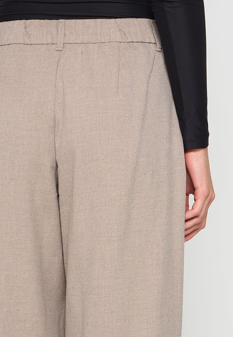 Nahaufnahme einer Person in beigen weiten Hosen mit Gürtelschlaufen und einem schwarzen Langarmoberteil, die Hand entspannt an ihrer Seite.