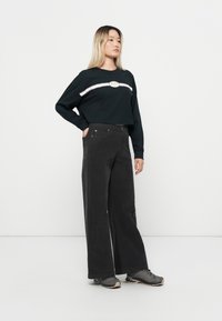Sweatshirt noir court avec une bande logo rayée blanche et bordeaux, associé à un pantalon large noir et des chaussures de sport grises.