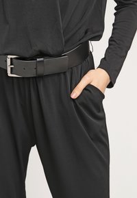 Combinaison noire avec une coupe décontractée, présentant une large taille, des poches profondes et une texture lisse et douce. Comprend une ceinture noire mate.