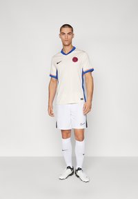 Vit fotbollströja med blå och orange accenter, matchad med vita shorts, vita strumpor och svarta och vita fotbollsskor. Nike-logotypen synlig.