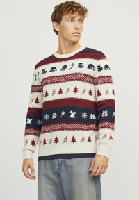 Pull de Noël tricoté arborant des couleurs rouge, marine et crème, avec des motifs d'arbres, de cadeaux et d'icônes saisonnières dans un design à rayures horizontales.