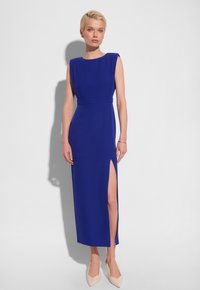 Królowska niebieska sukienka z bezrękawowym fasonem, boku rozcięciem, dopasowaną talią i gładką fakturą. Połączona z jasnobeżowymi butami na czubku.