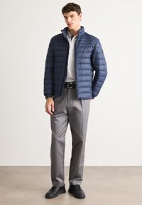 Man standing with one hand in pocket, wearing a half-zipped blue quilted jacket, white polo, gray pants, black belt, and black sneakers.