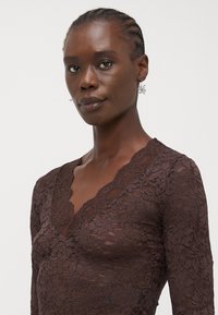 Brown lace top featuring a scalloped V-neckline and long sleeves. The lace has a floral pattern and a semi-transparent texture.