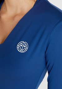 Modrý top s dlouhým rukávem a hlubokým výstřihem do V s bílým logem představujícím stylizovanou tvář na levé straně hrudi. Hladký povrch.