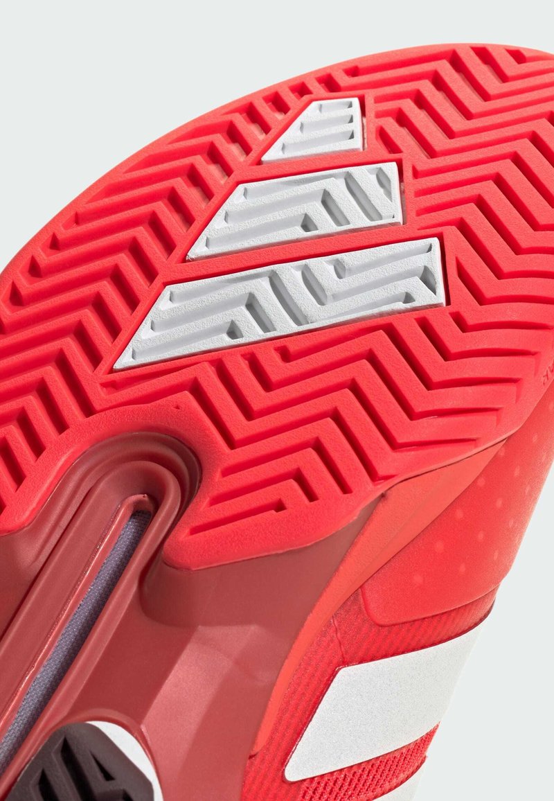 Semelle de chaussure de sport rouge avec caoutchouc texturé, trois rayures blanches, motif en zigzag et un design courbé prononcé.