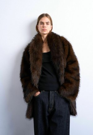 Jeune femme portant un manteau en fourrure marron foncé oversize, un haut noir et un jean foncé, debout les mains dans les poches devant un fond blanc.