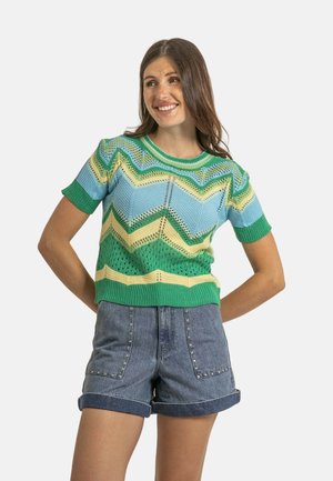 Pull en tricot à motif chevron bleu, vert et jaune, avec des manches courtes et un col côtelé vert, porté avec un short en jean.