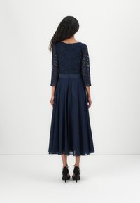 Robe bleu marine avec un corsage en dentelle et des manches trois-quarts ; jupe fluide avec une taille ajustée, accompagnée de chaussures à talons hauts noires.