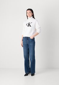 Hvit genser med høy hals og svart logo, kombinert med blå rette jeans og svarte spisse hæler, stående mot en nøytral bakgrunn.