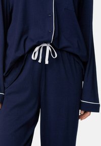 Mørkeblå pyjamas sæt med lange ærmer, hvid piping, en knaplukket top, og en justerbar hvid snøre i taljen på bukserne. Blødt stof med en behagelig tekstur.