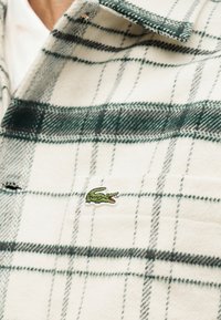 Κοντινό πλάνο λευκού καρό πουκαμίσου με πράσινες γραμμές, με μικρό κεντημένο πράσινο λογότυπο κροκόδειλου στην περιοχή της τσέπης στο στήθος.