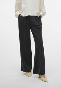 Zwarte wijde broek van soepel materiaal met plooien, gecombineerd met een witte blouse met kantaccenten en lange mouwen.
