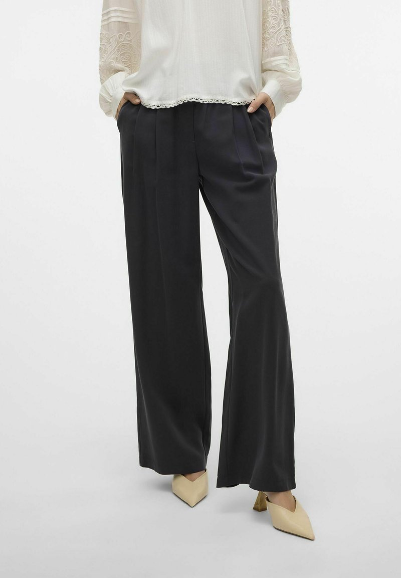 Zwarte wijde broek van soepel materiaal met plooien, gecombineerd met een witte blouse met kantaccenten en lange mouwen.