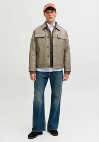 Olivgrüne Lederjacke mit zwei Brusttaschen, über einem braunen Strickpullover, weißem Hemd, blauen Schlagjeans und schwarzen Schuhen getragen.