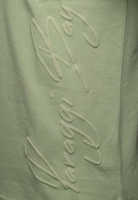 Vaaleanvihreä huppari, jossa on kohokuvioitu teksti. Kangas on teksturoitu pehmeällä pinnalla, mikä korostaa hienoa väriä.