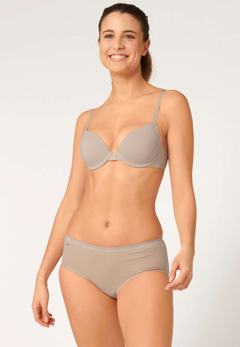 Femme souriante aux cheveux bruns attachés en queue de cheval, portant un soutien-gorge beige assorti à sa lingerie, debout devant un fond clair uni.
