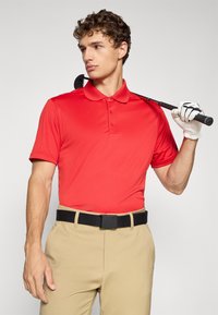 Mladý muž v červenej polo košeli a béžových nohaviciach drží golfovú palicu cez rameno, na pravej ruke má bielu golfovú rukavicu.