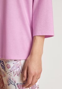 Korte mouwen roze top van zachte stof, met een ronde halslijn. De hand die te zien is, steunt op een patroonbroek in lichte kleur met bloemenmotieven.