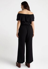 Svart off-shoulder jumpsuit med volangöverdel, elastisk midja, vida ben och plattformsskor med nitar. Slät textur.