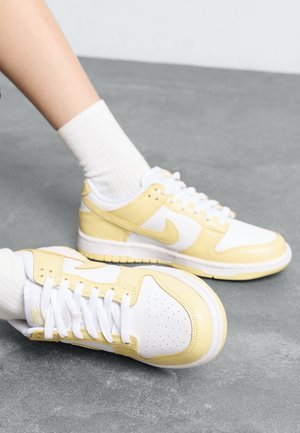 Tenisky Nike s bílou a žlutou barevnou kombinací, koženým a síťovaným materiálem, perforovanou špičkou a klasickým designem na šněrování.
