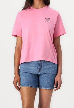 Femme portant un t-shirt rose à manches courtes avec un petit imprimé floral et un short en denim bleu sur fond blanc.