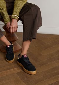 Black suede platform sneakers with a textured pattern, dark laces, a burgundy accent on the heel, and a thick cork sole. Brown trousers are worn.