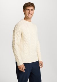 Fester cremefarbener Kabelstrickpullover mit strukturierten Mustern, gerippten Bündchen und Saum, getragen über dunkelblauen Hosen. Leichte Strickmaterialien.