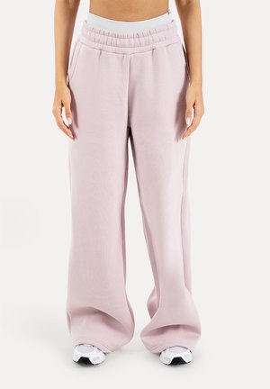 Frau trägt hoch taillierte, weit geschnittene hellrosa Sweatpants mit elastischem Bund und weißen Sneakern, steht vor einfachem Hintergrund.