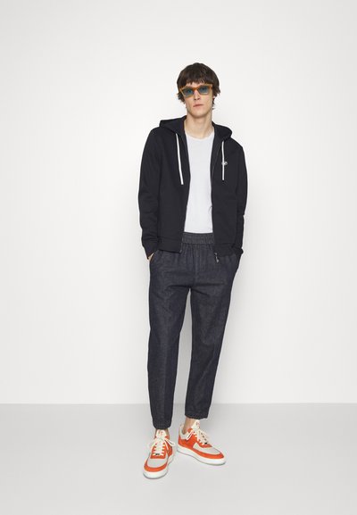 Black zip-up hoodie over a white t-shirt, dark denim tapered pants with elastic cuffs, and orange and white sneakers. Light blue sunglasses.