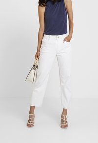Jean amplio de mezclilla blanca, combinado con una blusa sin mangas azul marino y tacones de tiras color beige. Se sostiene un pequeño bolso crema con una mano.