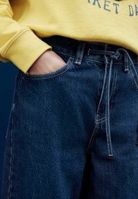 Persona, kas valkā tumši zilas džinsa bikses ar saišu jostu un dzeltenu sporta džemperi, roka iegriezta kabatā uz tumša fona.