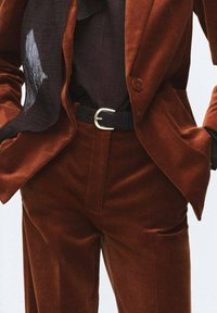 Herenkostuum van bruin fluweel met een getailleerde blazer en op maat gemaakte broek. Zwarte riem met een zilveren gesp. Doorzichtige zwarte sjaal die om de hals is gedrapeerd.