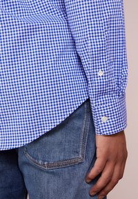 Camicia a quadri blu e bianchi con polsini abbottonati, abbinata a jeans in denim blu. La camicia presenta un orlo curvo e una texture liscia.