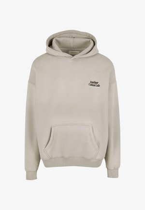 Ljusbeige hoodie med ficka framtill och liten svart text "Another Cotton Lab" på vänster bröst.